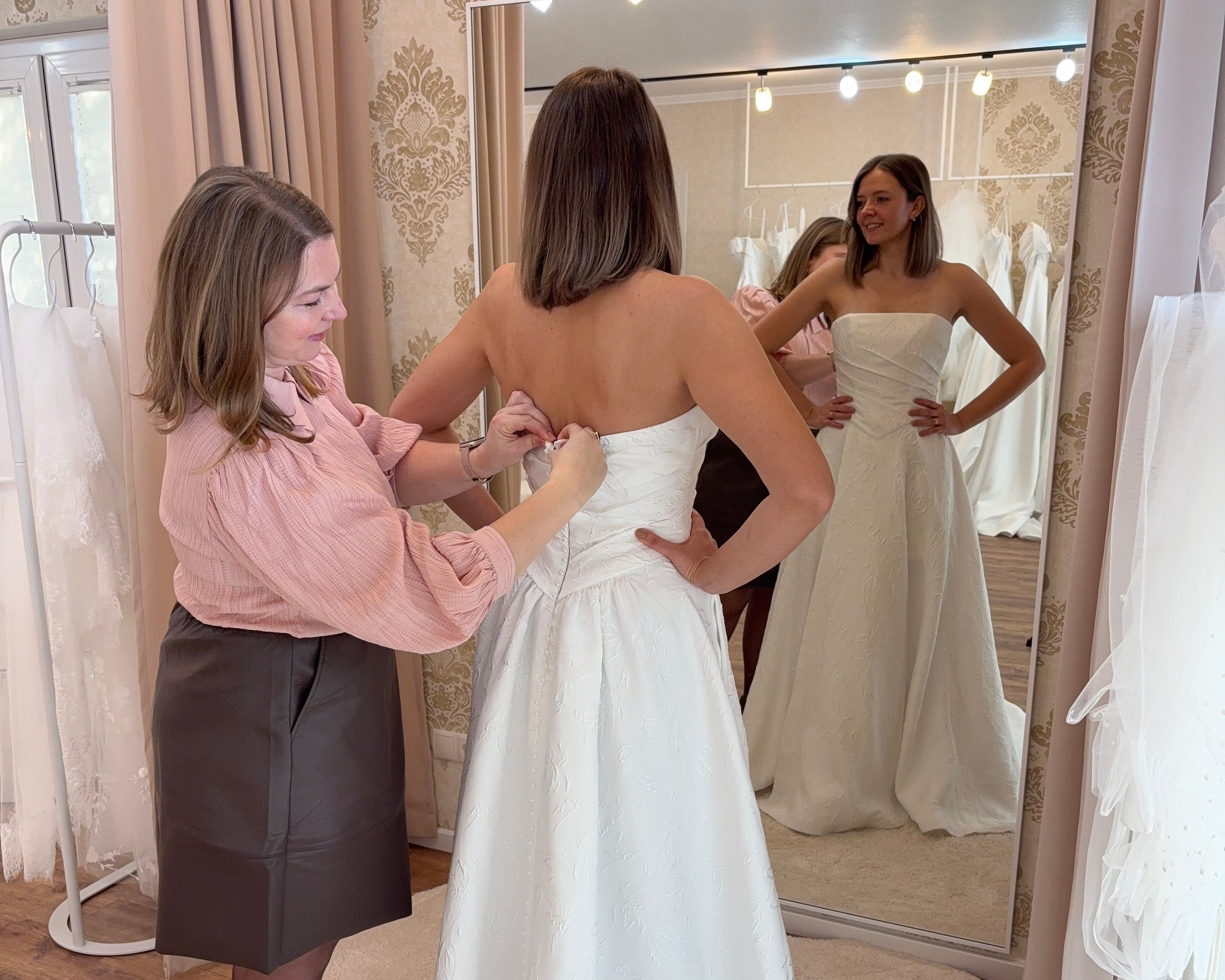 Brautmode Wuerzburg: Blick in die Anprobe. Brautstylistin knöpft den Brautkleid an einer Braut.