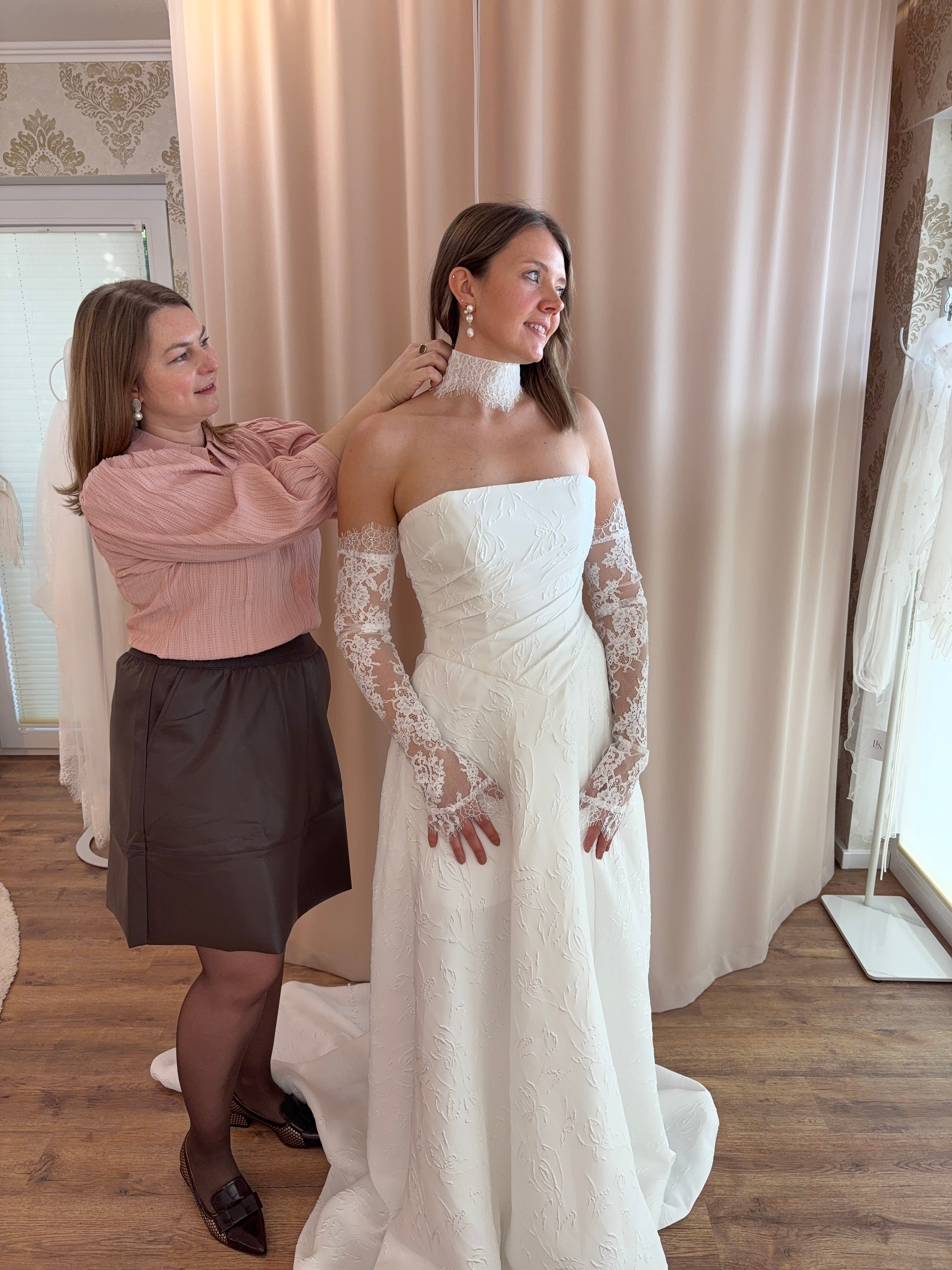 Brautmode Würzburg – Anprobe in der Boutique: Beraterin richtet einer Braut das schulterfreie Brautkleid vor dem Spiegel.