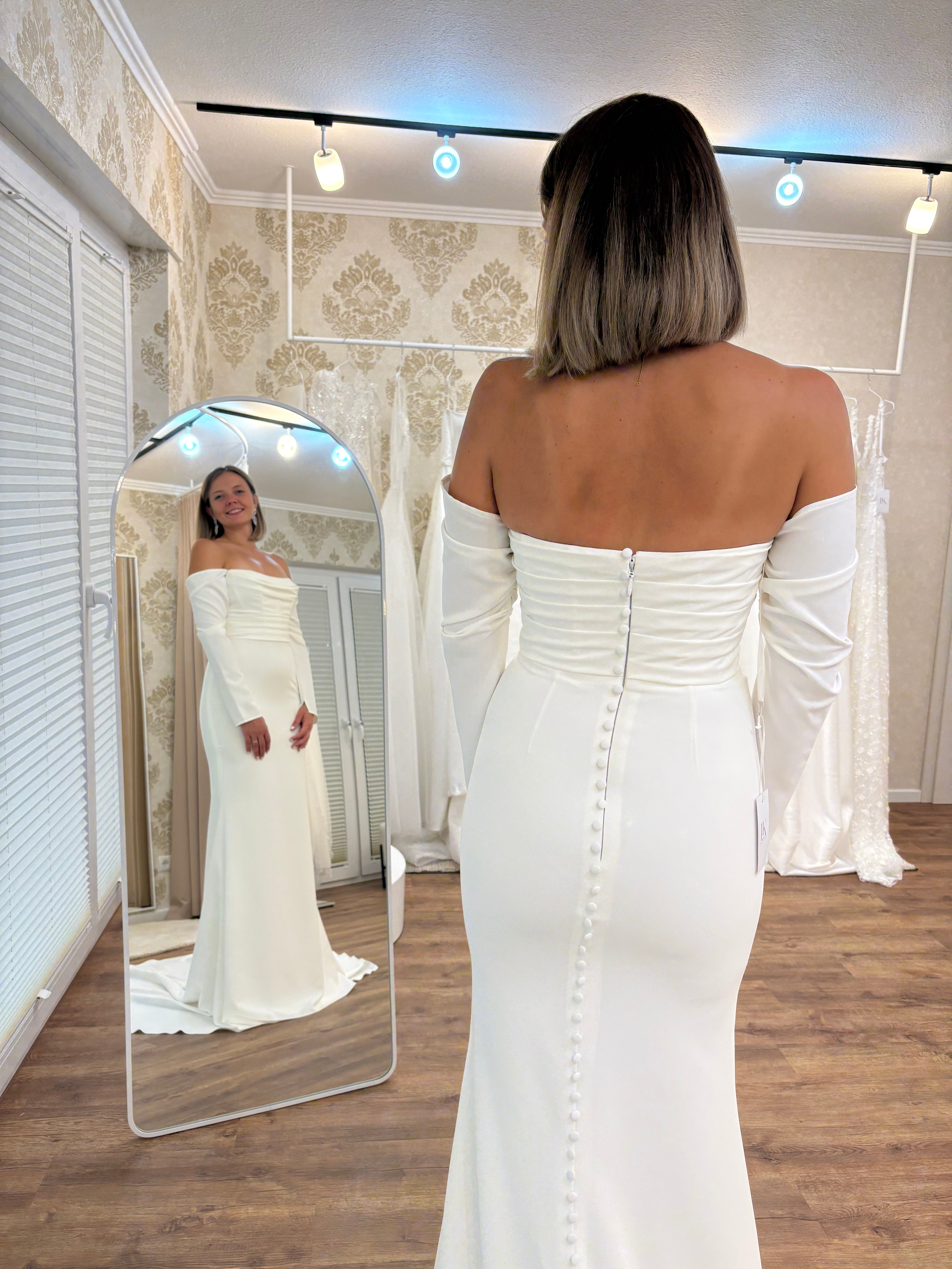 Brautmode Würzburg – Braut im schlichten, figurbetonten Brautkleid mit Knopfleiste am Rücken vor einem Spiegel in der Boutique.