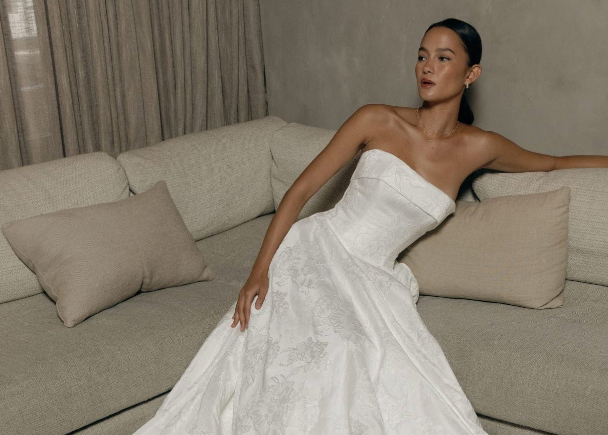 Brautmode Würzburg – Braut in trägerlosem Brautkleid mit floraler Struktur, elegant auf einem Sofa sitzend.