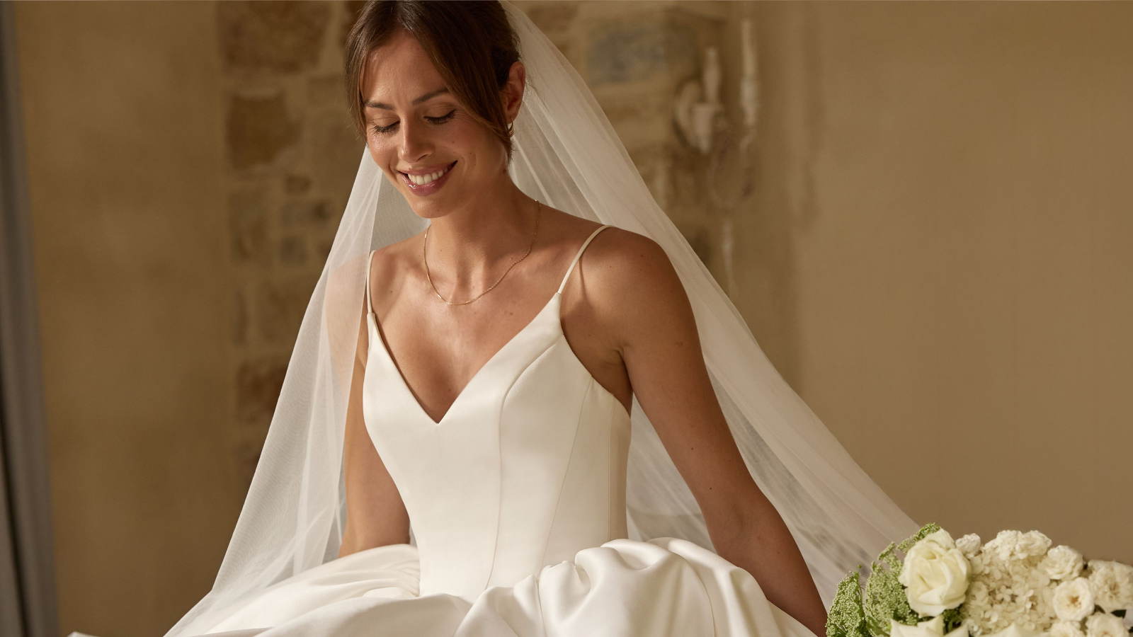 Brautmode Würzburg – lächelnde Braut im schlichten V-Ausschnitt-Brautkleid mit Schleier in warmer Naturkulisse.